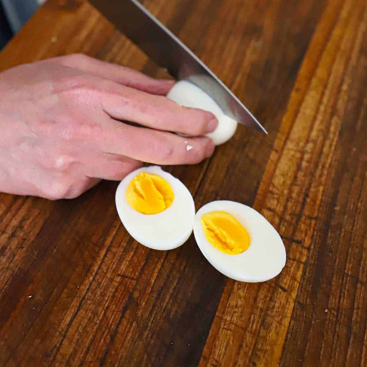 A person using a chef's knife to slice a hard boiled egg in half on a cutting board.