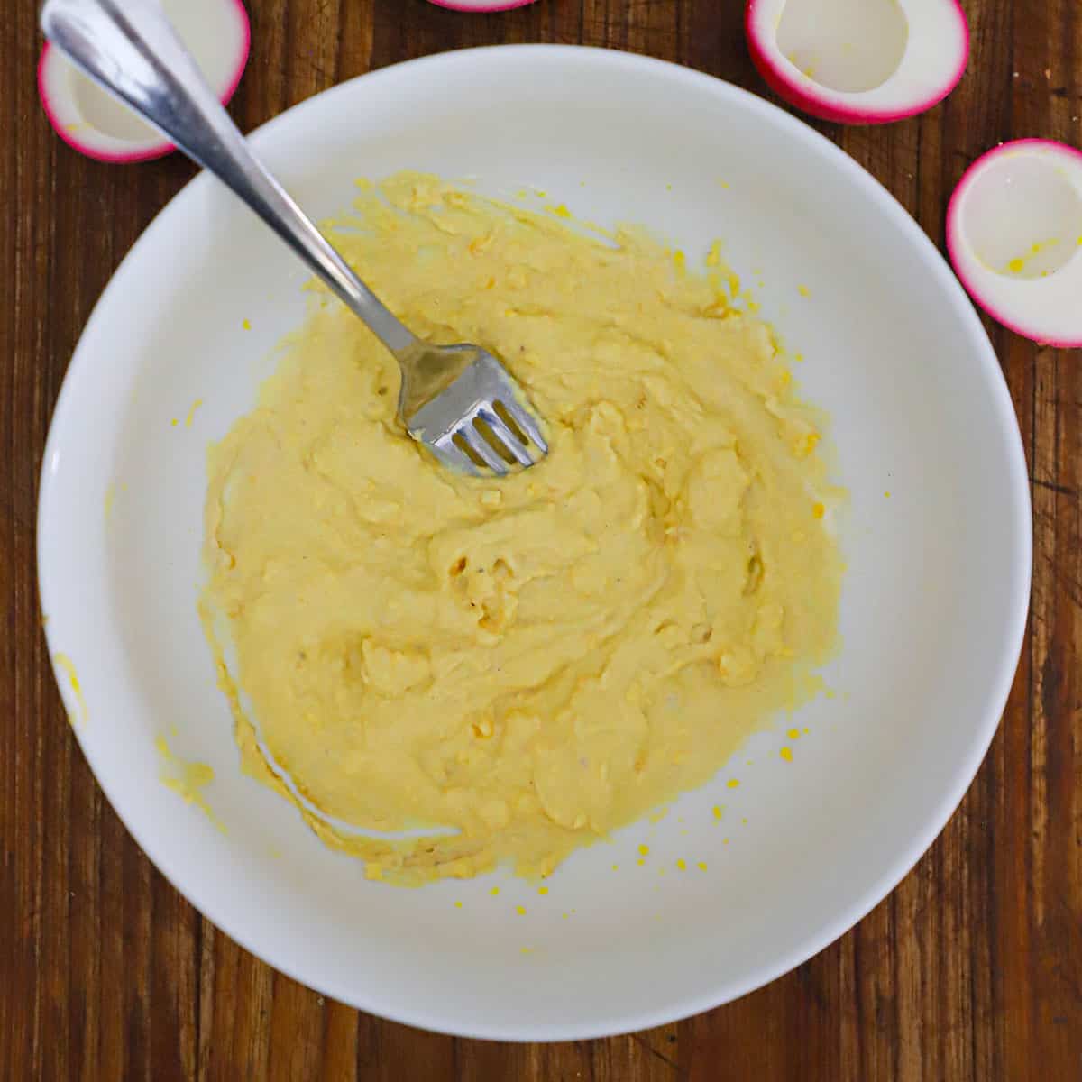 A person using the back of a fork to mash the filling for horseradish beet deviled eggs in a white bowl.