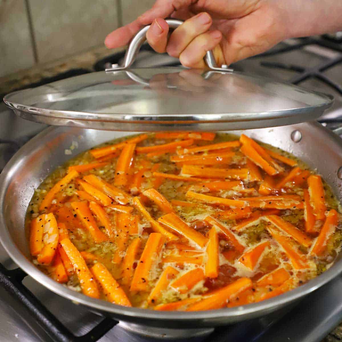 A person lifting a glass lid from a skillet filled with simmering maple-glazed carrots.