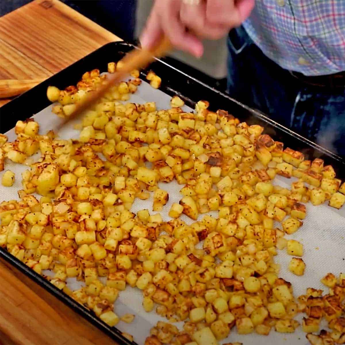 A person using a wooden spatula to move around crispy cubed breakfast potatoes on a baking sheet pan.