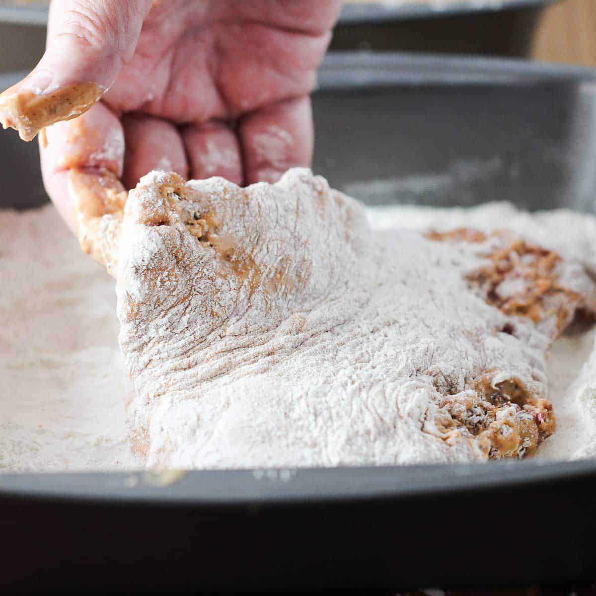 A person dredging a cube steak that has been dipped into a wet batter through seasoned flour in a square pan.