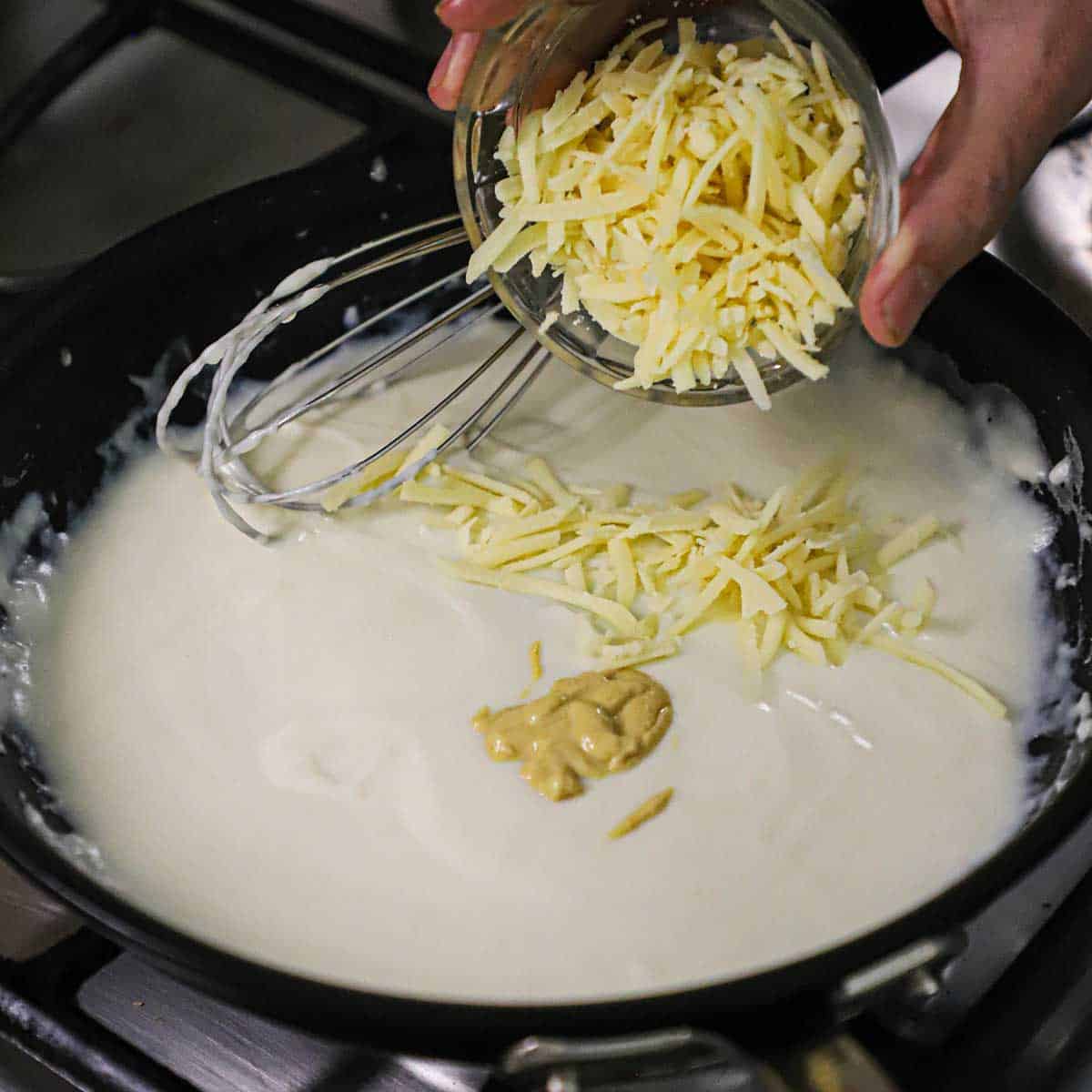 A person dumping grated Gruyére cheese into a skillet filled with a béchamel sauce.