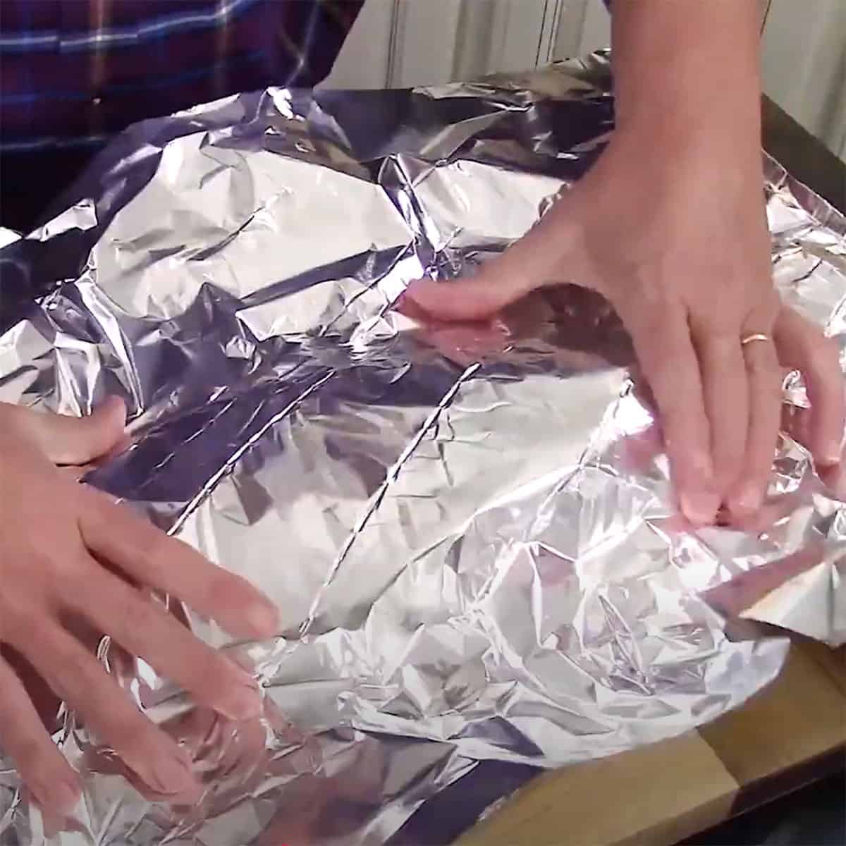 A person placing a large piece of aluminum foil over a large cooked brisket on a wooden cutting board.