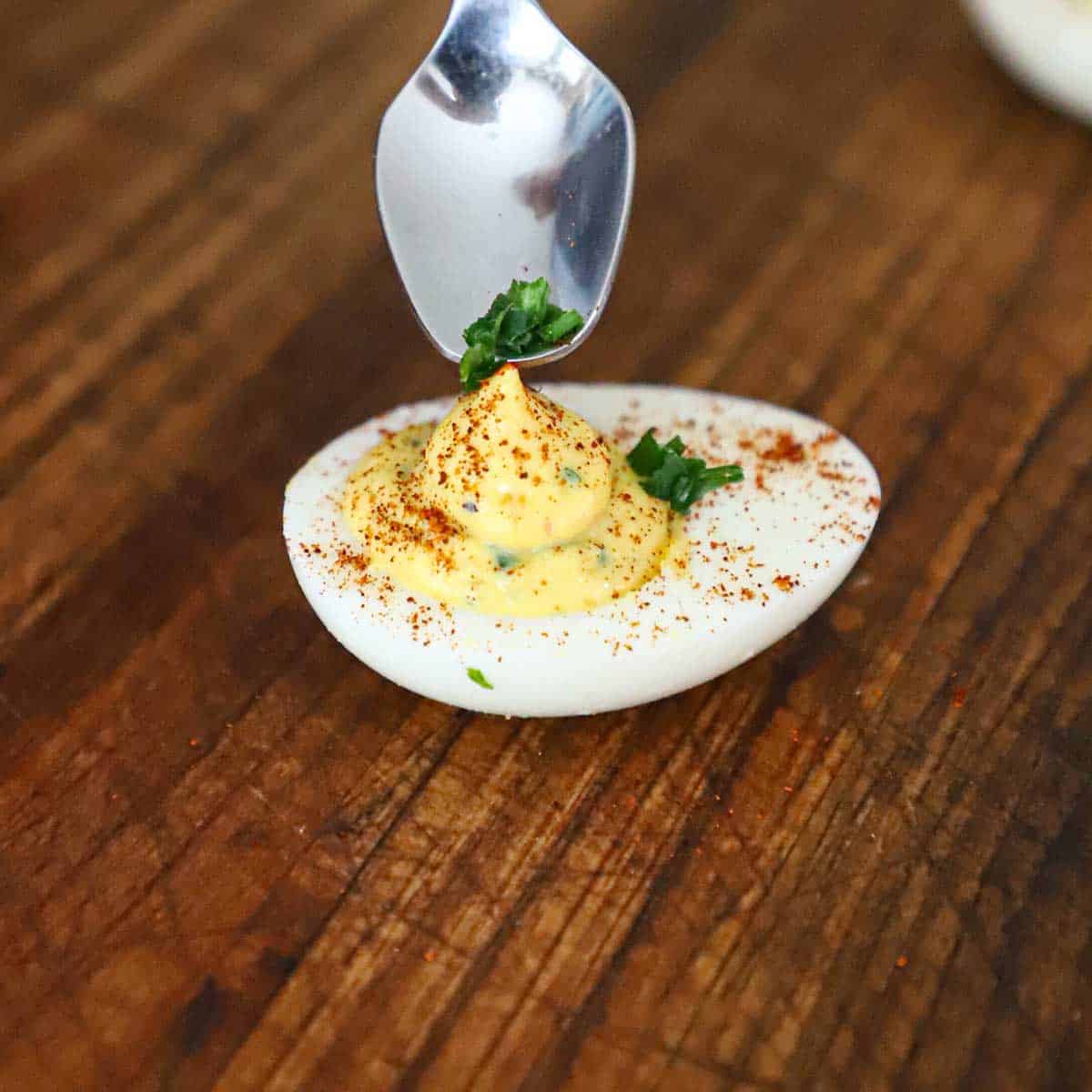 A person using a small spoon to place snipped chives on the top of a traditional deviled egg.