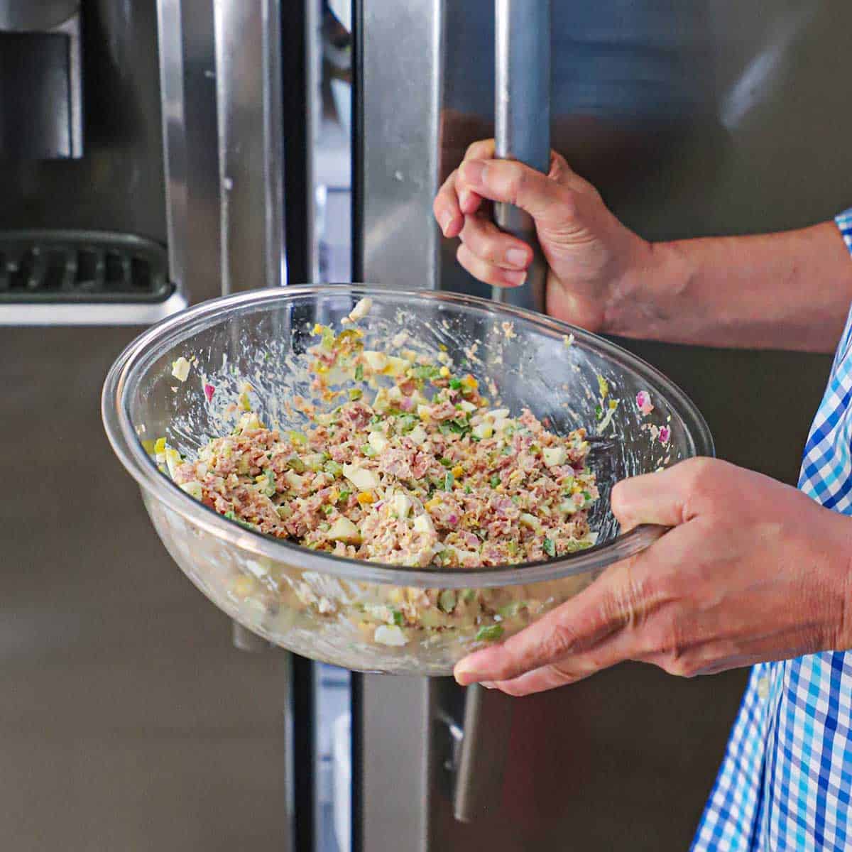 A person using one hand to hold a large glass bowl filled with best-ever ham salad and the other hand on the door handle of the refrigerator that he is getting ready to open.
