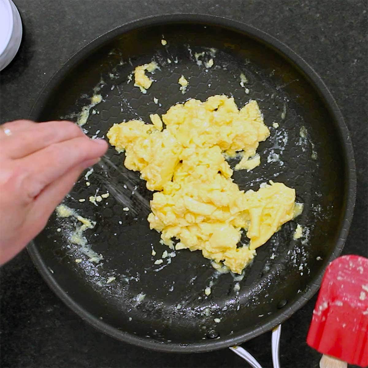 A person sprinkling salt onto a pile of freshly prepared fluffy scrambled eggs in a black non-stick skillet.