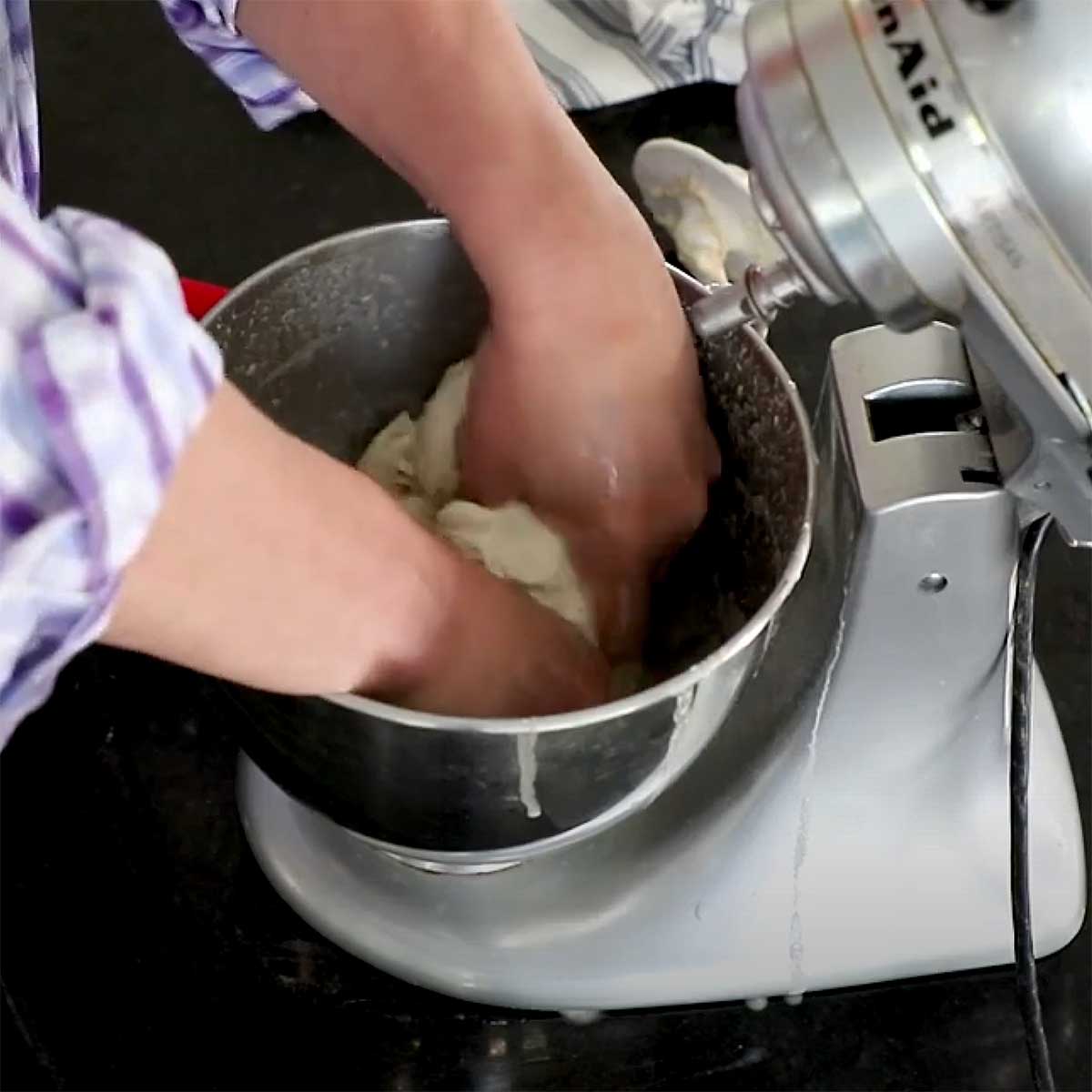 A person using both hands to massage a yeast mixture into a ball of bread dough in the bowl of a stand mixer.