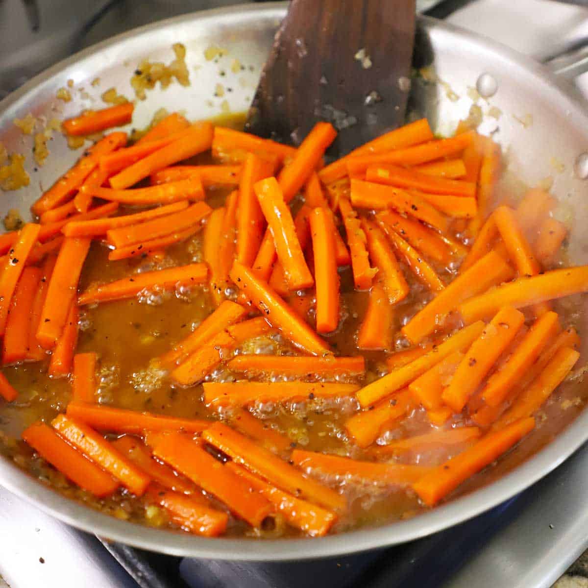 A person using a wooden spatula to stir maple-glazed carrots in a large silver skillet.