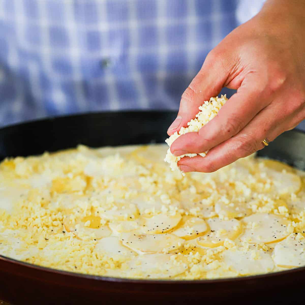 A person sprinkling grated Gruyere cheese over a baking dish filled with uncooked potatoes dauphinoise.