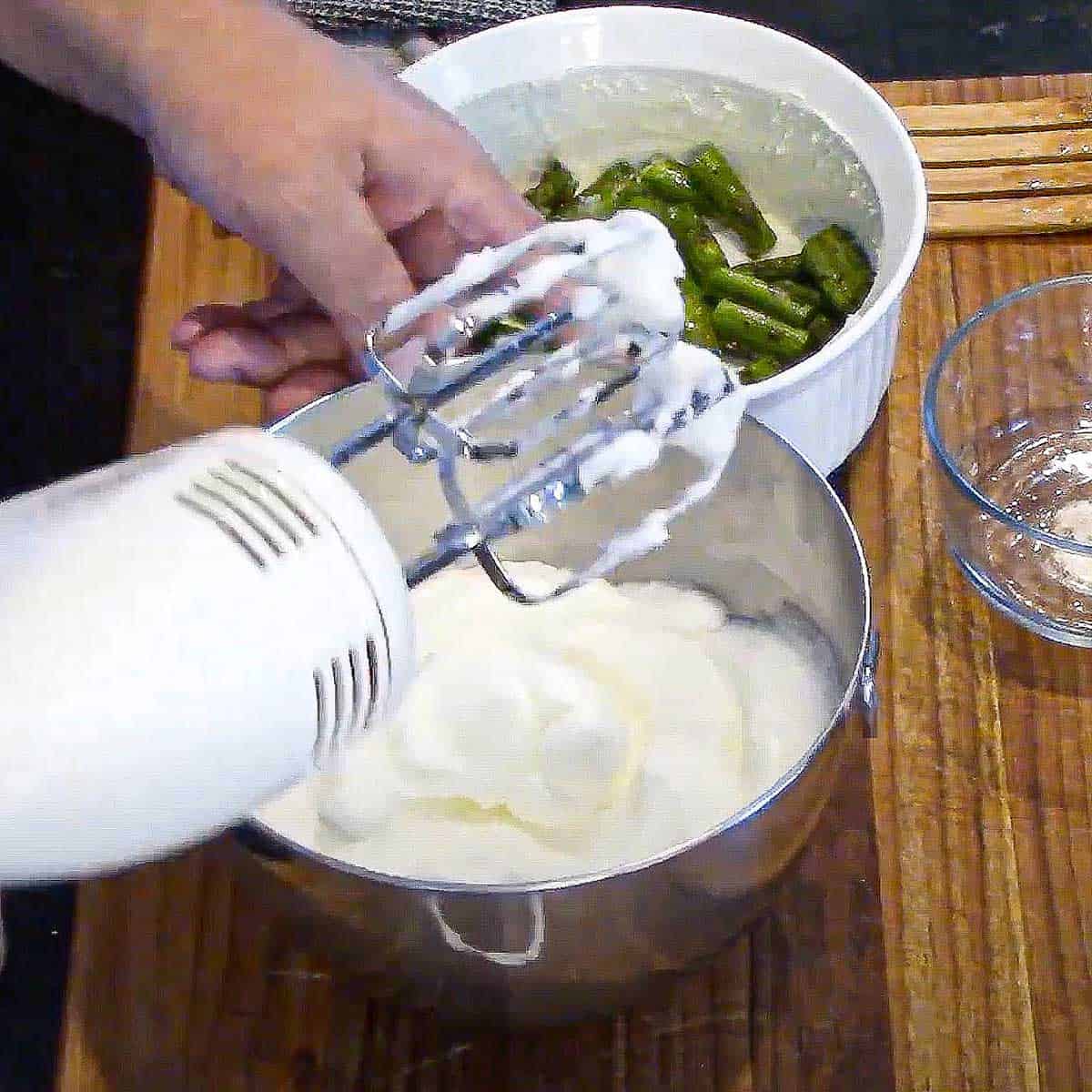 A person holding up a hand-held electric mixer over a bowl of egg whites that have been beaten into soft peaks.