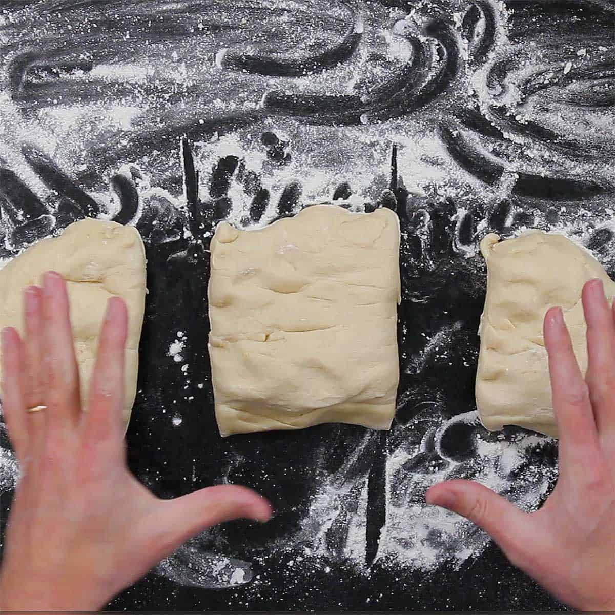 A person who a cut dinner roll dough into thirds on a floured black marble surface.