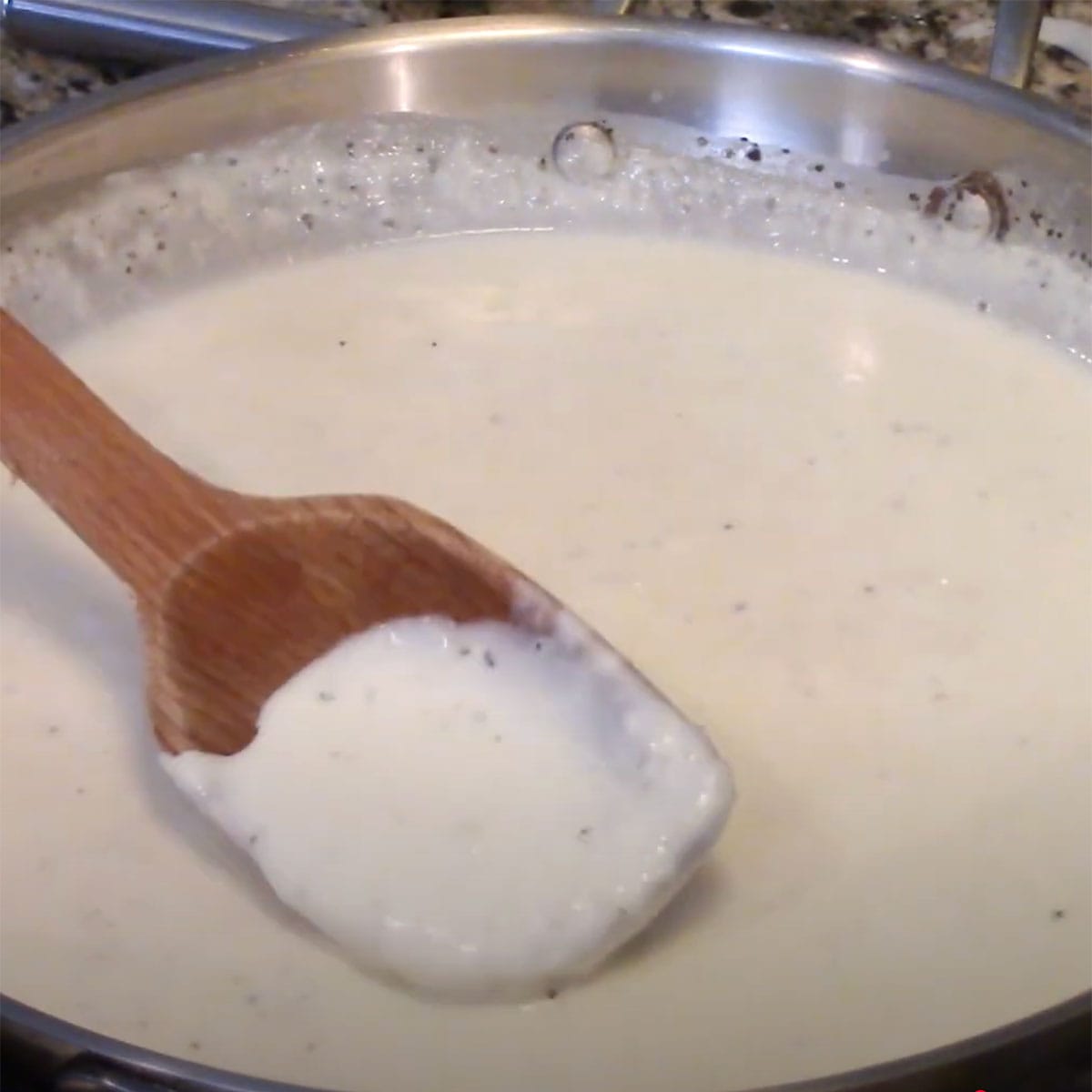 A person stirring a wooden spoon into a thickened cream gravy in a large silver skillet.