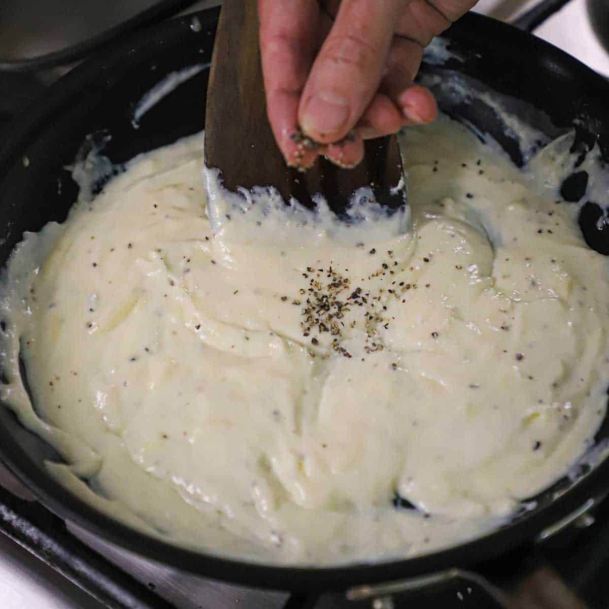 A person using his finger to sprinkle black pepper into a skillet filled with a Mornay sauce.