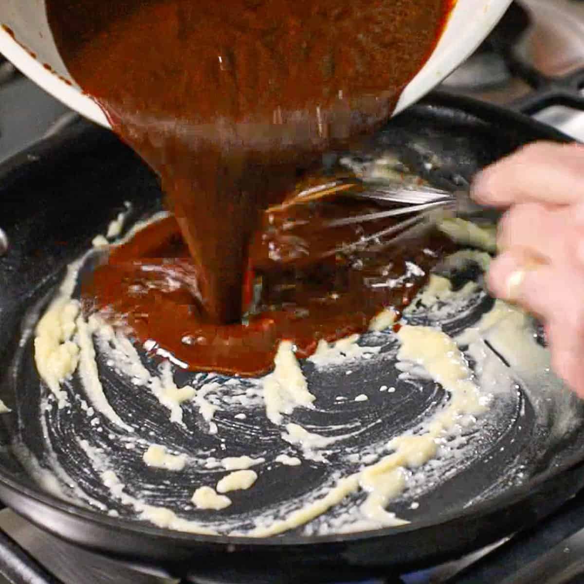 A person pouring a puréed chile and vegetables sauce into a skillet with a roux simmering in it.