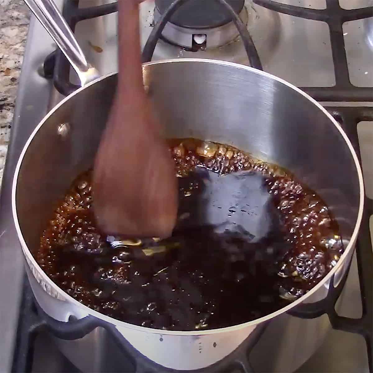 A person using a wooden spoon to stir a simmering au jus sauce in a silver saucepan on a stove.