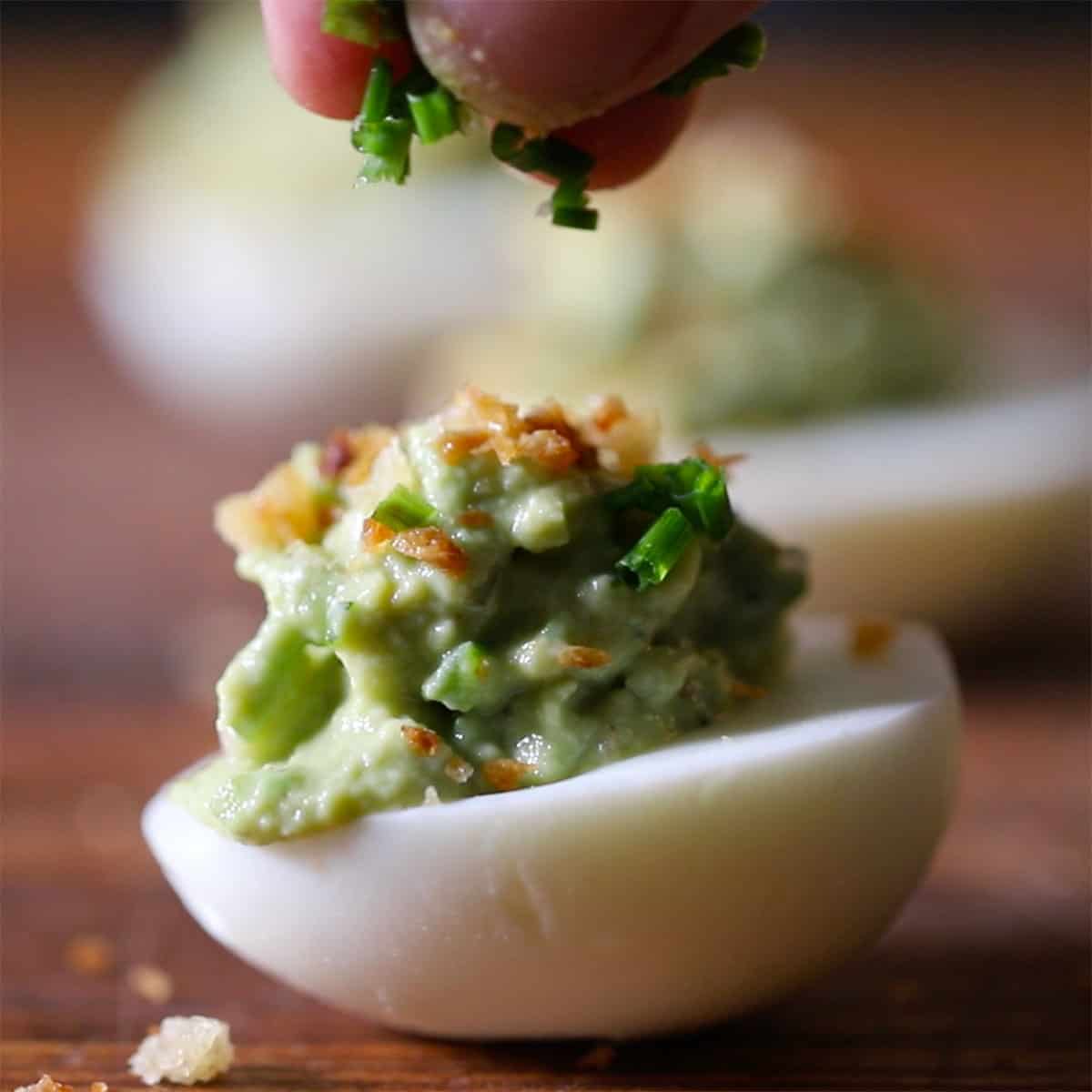 A person sprinkling snipped chives over the top of an avocado deviled egg sitting o a wooden cutting board.