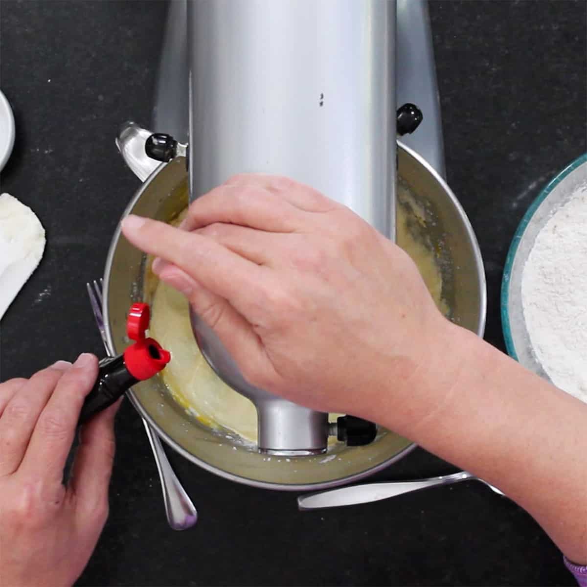 A person pouring almond extract from a small plastic container into the bowl of a stand mixer filled with creamed butter and sugar.