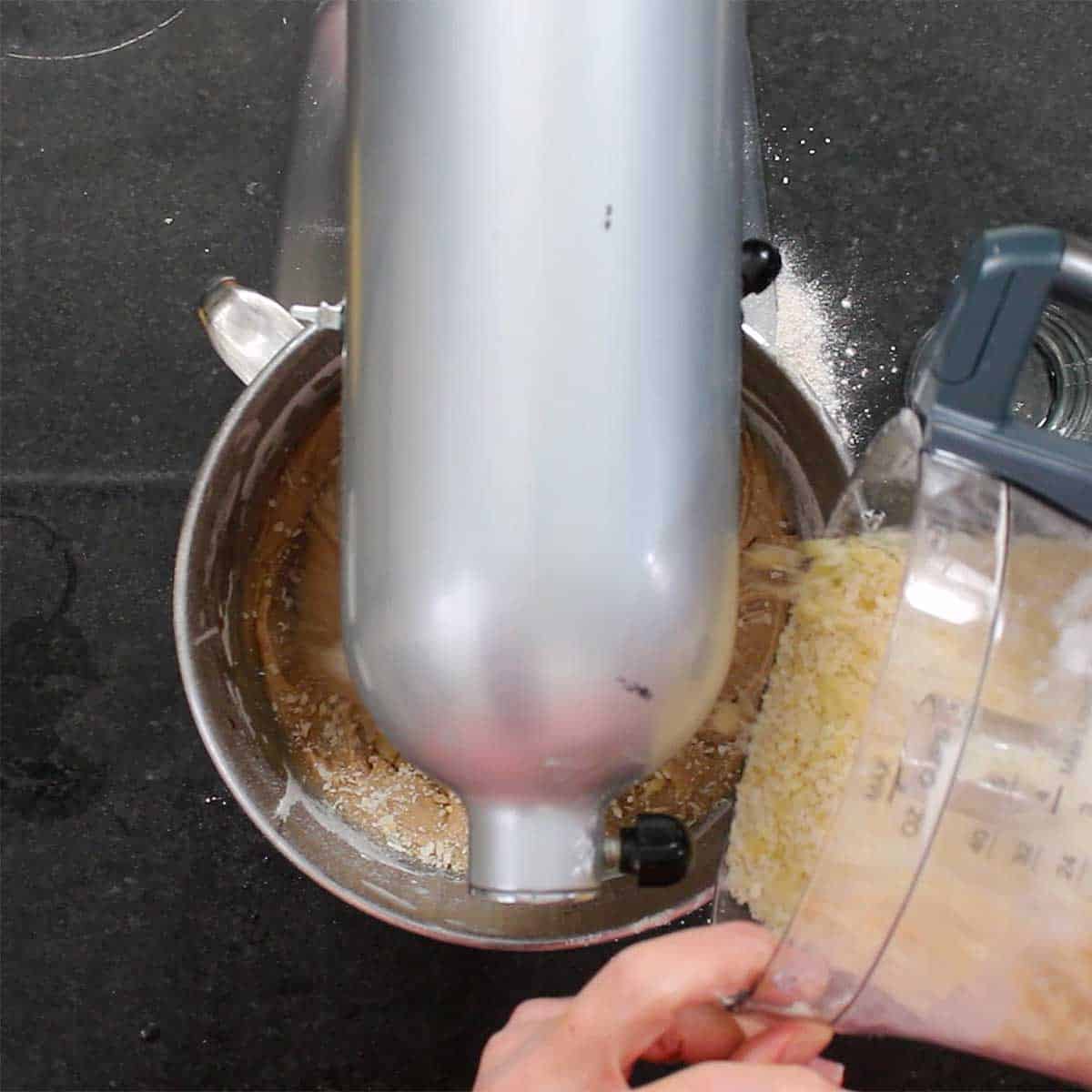 A person adding toasted coconut into the bowl of a stand mixer.