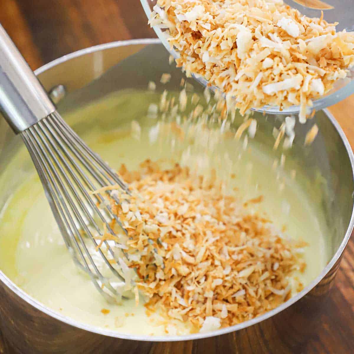 A person pouring toasted coconut flakes from a glass bowl into a saucepan with custard that is being stirred with a whisk.