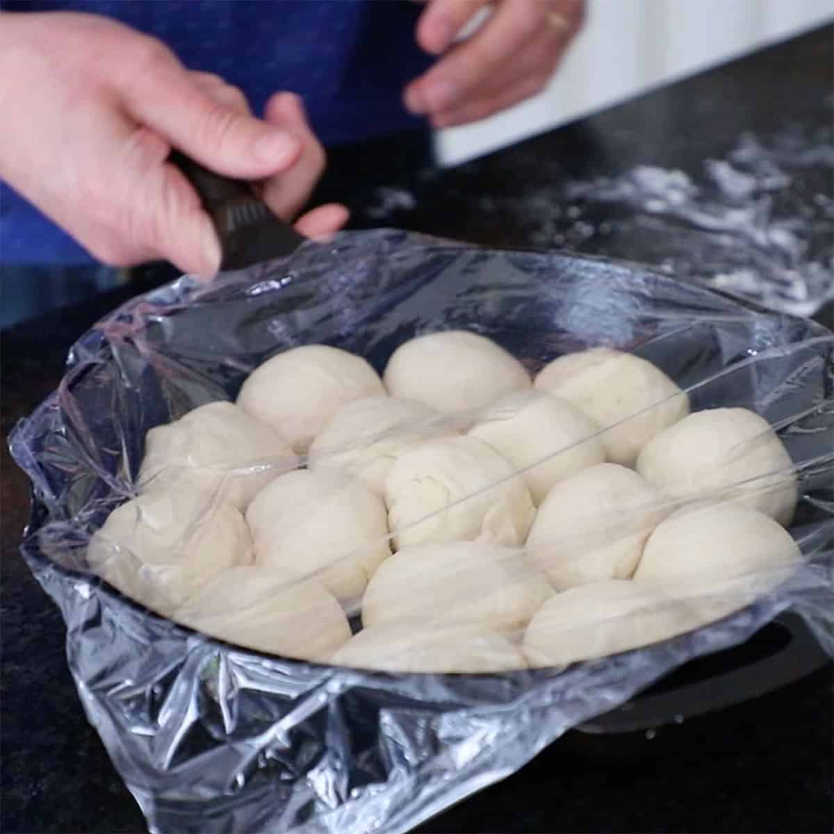 A person picking up a cast-iron skillet that is filled with round balls of dough and is covered with plastic wrap.