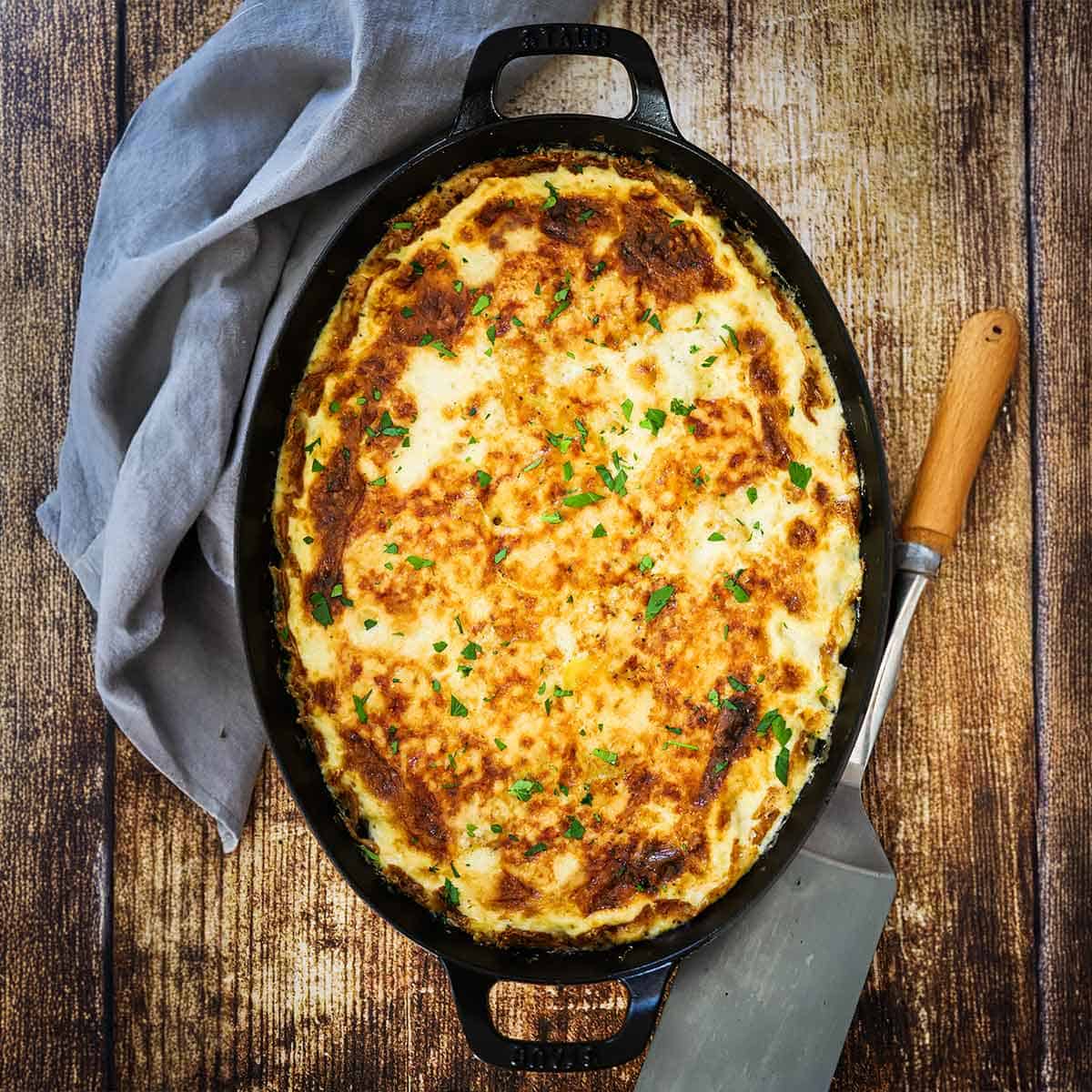 A fully cooked dish of potatoes dauphinoise that is resting on a wooden background with a large spatula and grey napkin nearby.