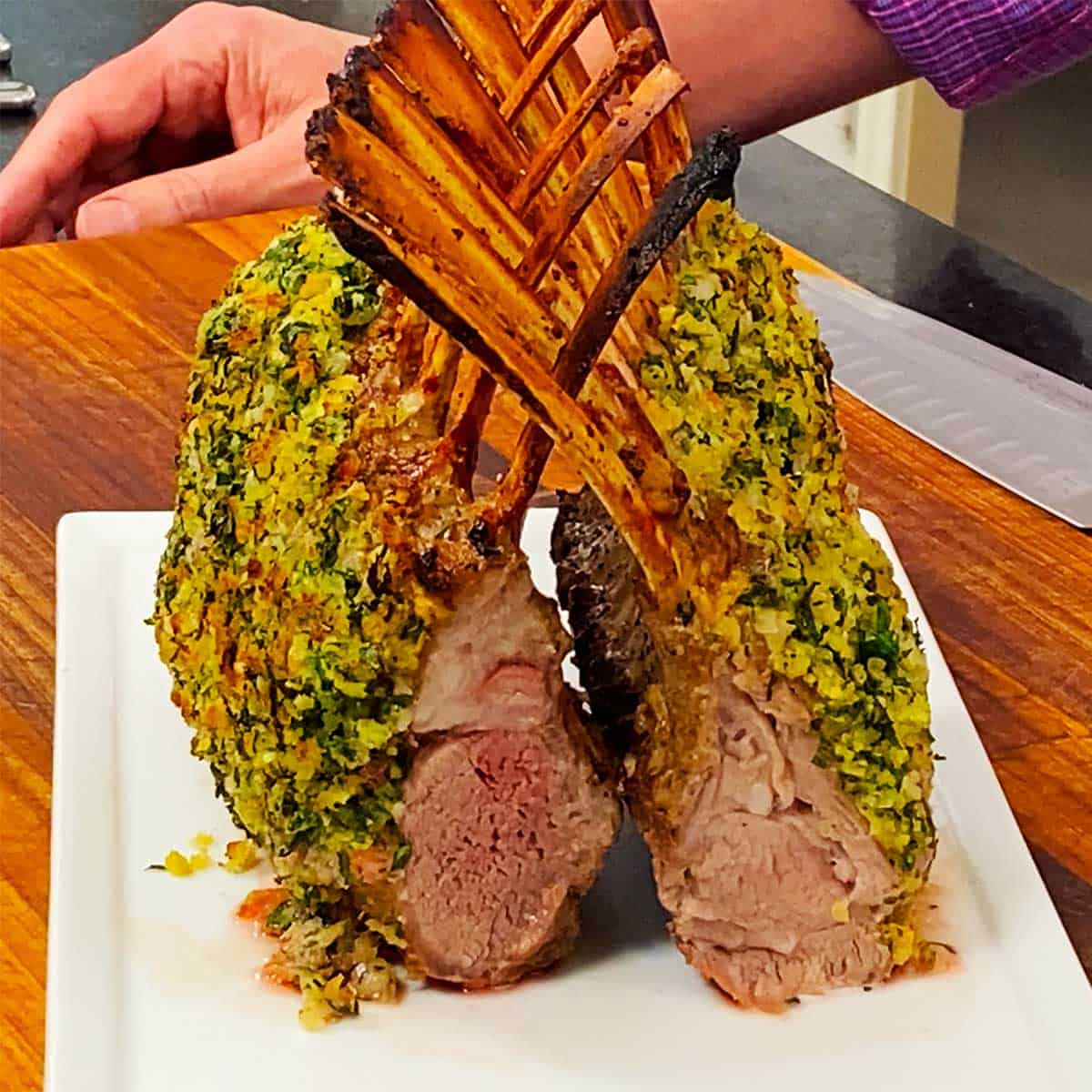 A person standing behind a platter that is holding a pair of roast rack of lamb persillade that are standing upright with their bones intersecting each other in a "cathedral" style presentation.