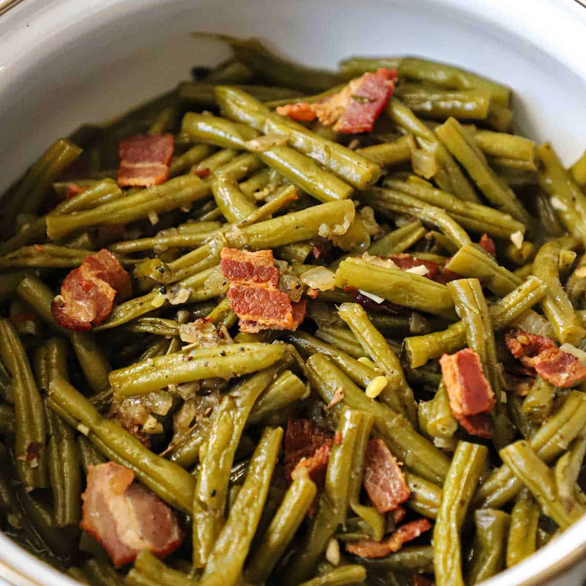 A white serving bowl filled with Southern-style green beans that have pieces of crispy bacon on top.