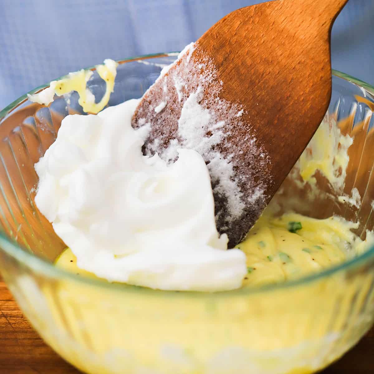 A person using a wooden spatula to fold fluffy egg whites into a soufflé batter in a glass bowl.