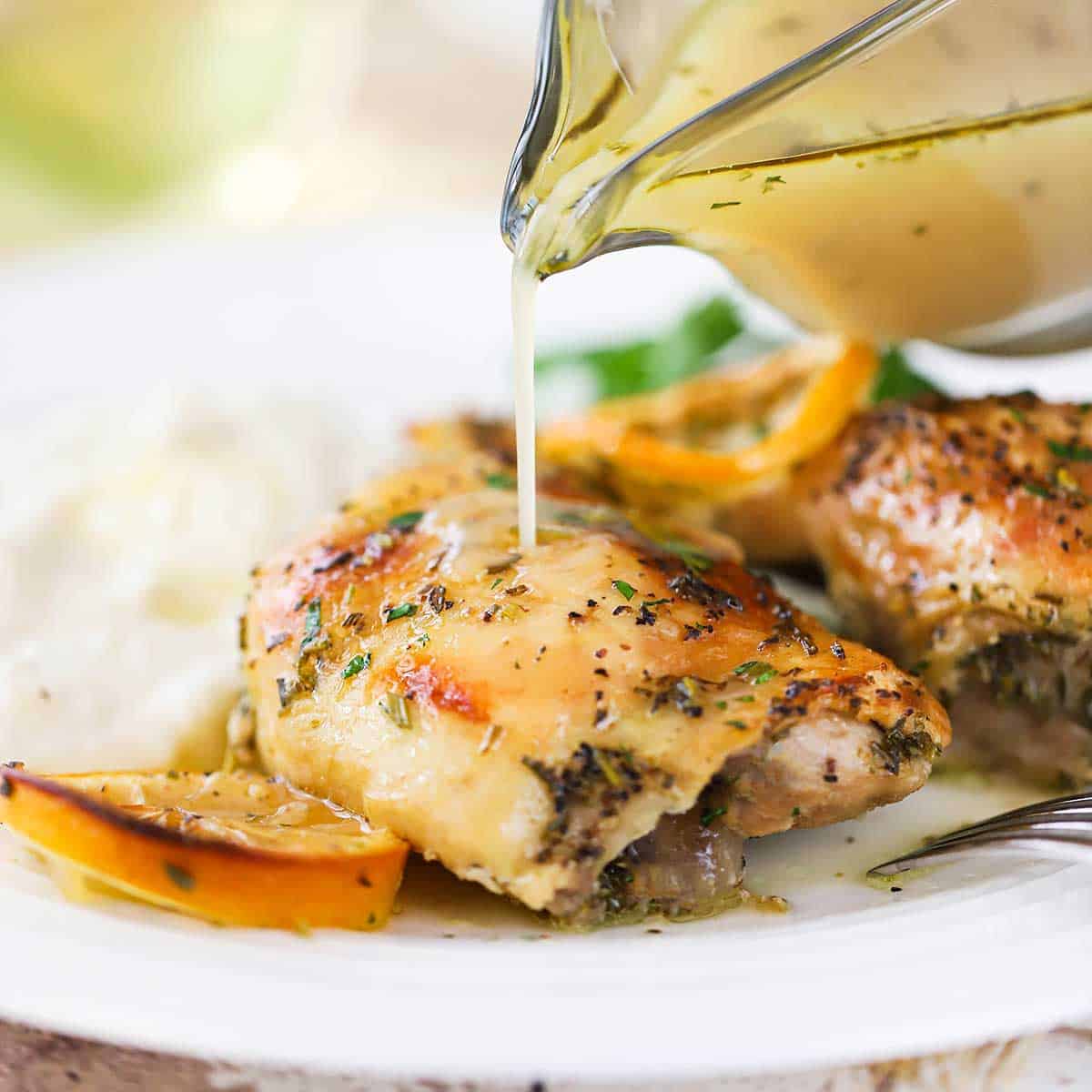 Lemon white wine sauce being poured from a glass gravy boat over piece of lemon roast chicken on a white dinner plate.