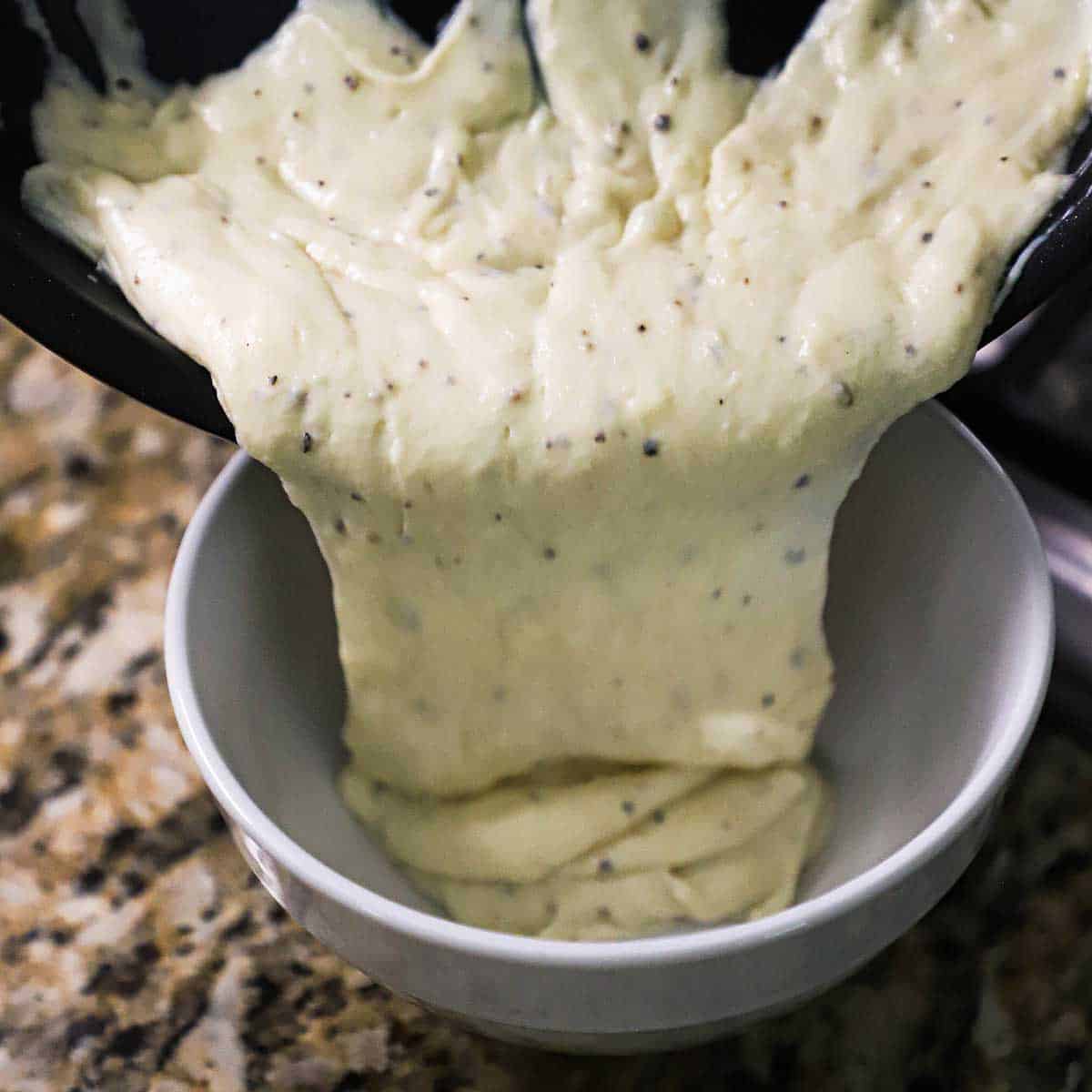 Mornay sauce being transferred from a skillet into a white ceramic bowl.