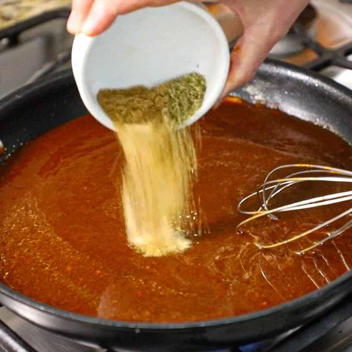 A person dumping seasonings into a skillet filled with a red enchilada sauce.