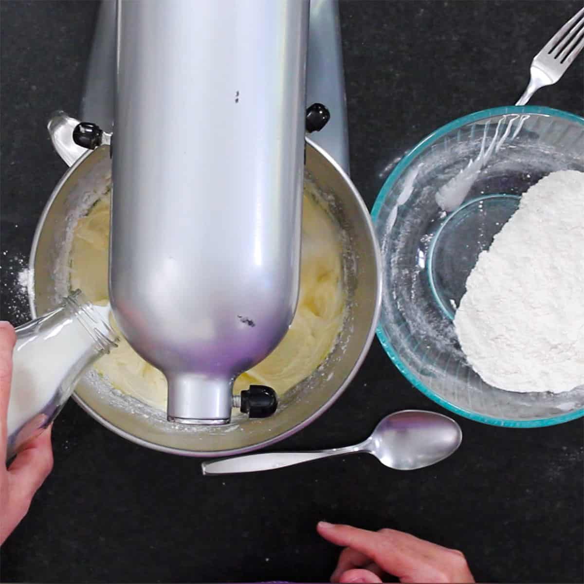 A person pouring buttermilk from a small glass jug into the bowl of a stand mixer with a glass bowl of a flour mixture nearby.
