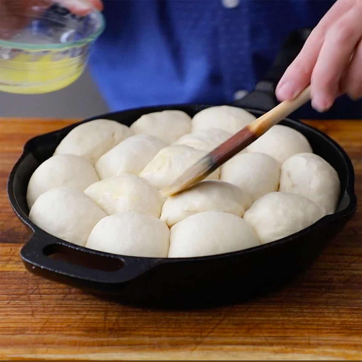 A person using a small pastry brush to brush melted butter over the tops of un-cooked dinner rolls in a cast-iron skillet.