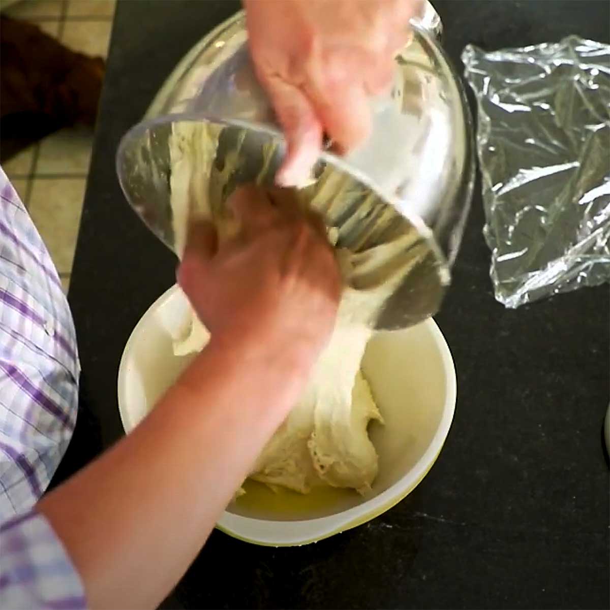 A person transferring a sticky bread dough from a mixing bowl into a ceramic bowl that has been greased with olive oil.