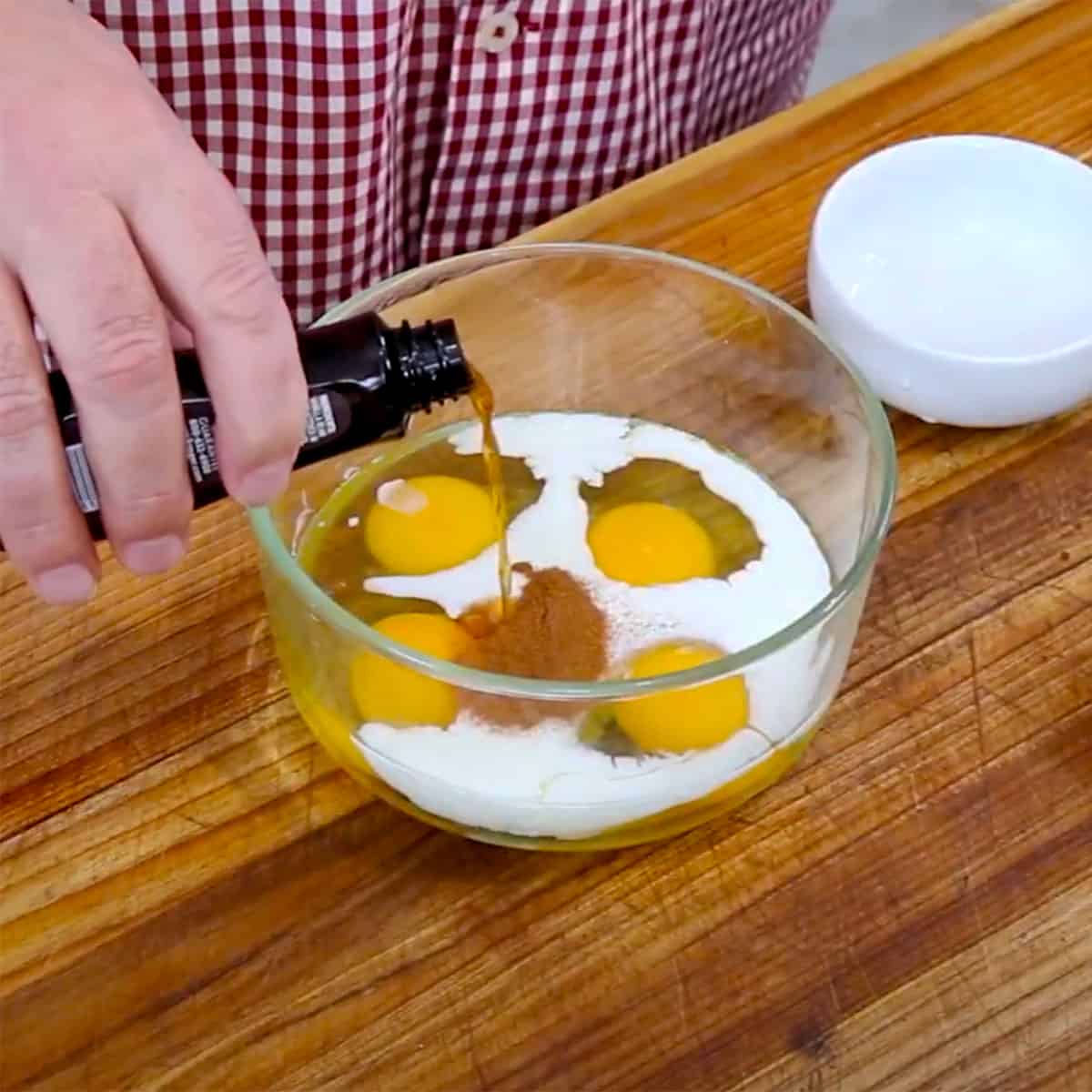 A person pouring vanilla extract from a small bottle into a glass bowl filled with whole eggs, milk, and ground cinnamon.