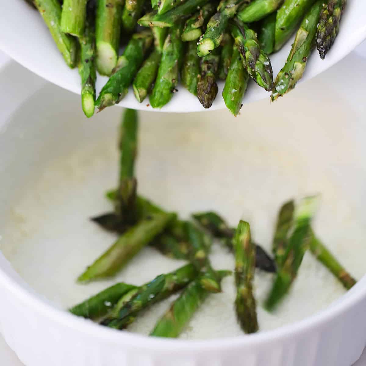 Roasted pieces of asparagus being dumped from a bowl into a soufflé dish that has been greased with butter and dusted with grated Parmesan cheese.