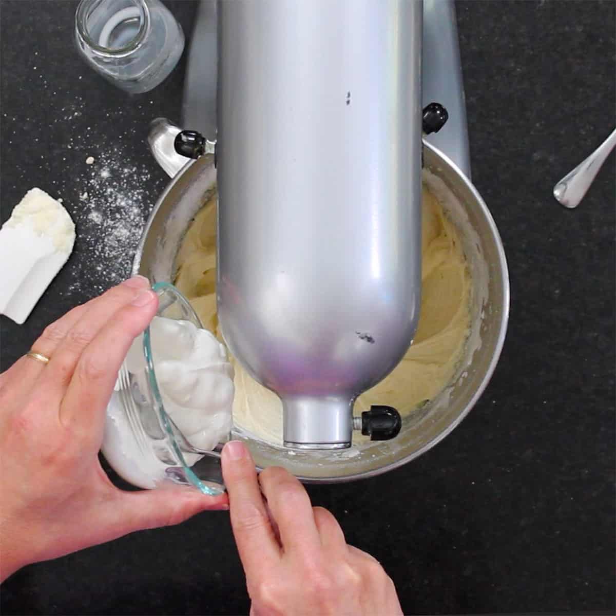 A person transferring sour cream from a small glass bowl into a stand mixer filled with coffee cake batter.