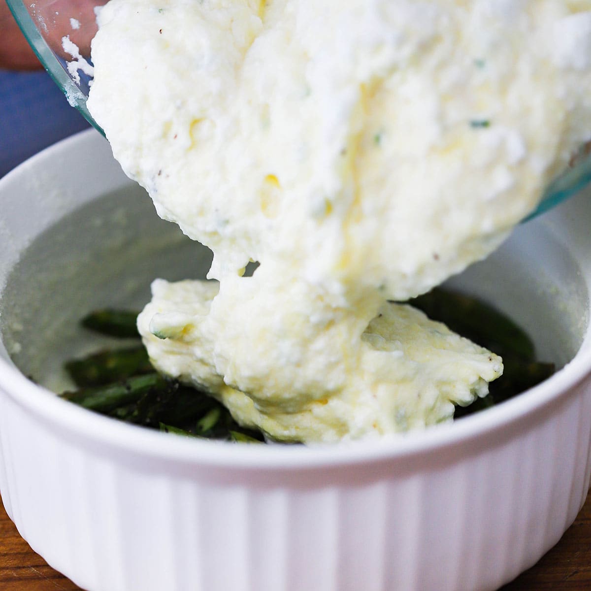 Soufflé batter being transferred from a glass bowl into a white circular baking dish that has roasted asparagus in the bottom of it.