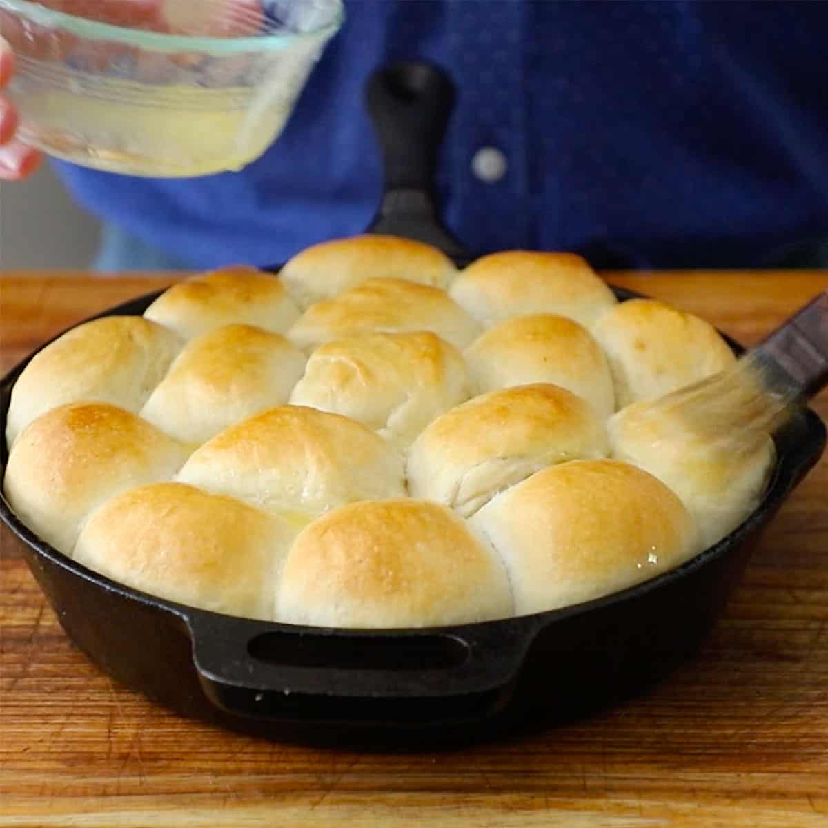 A person brushing melted butter over the tops of freshly baked skillet dinner rolls in a cast-iron skillet.