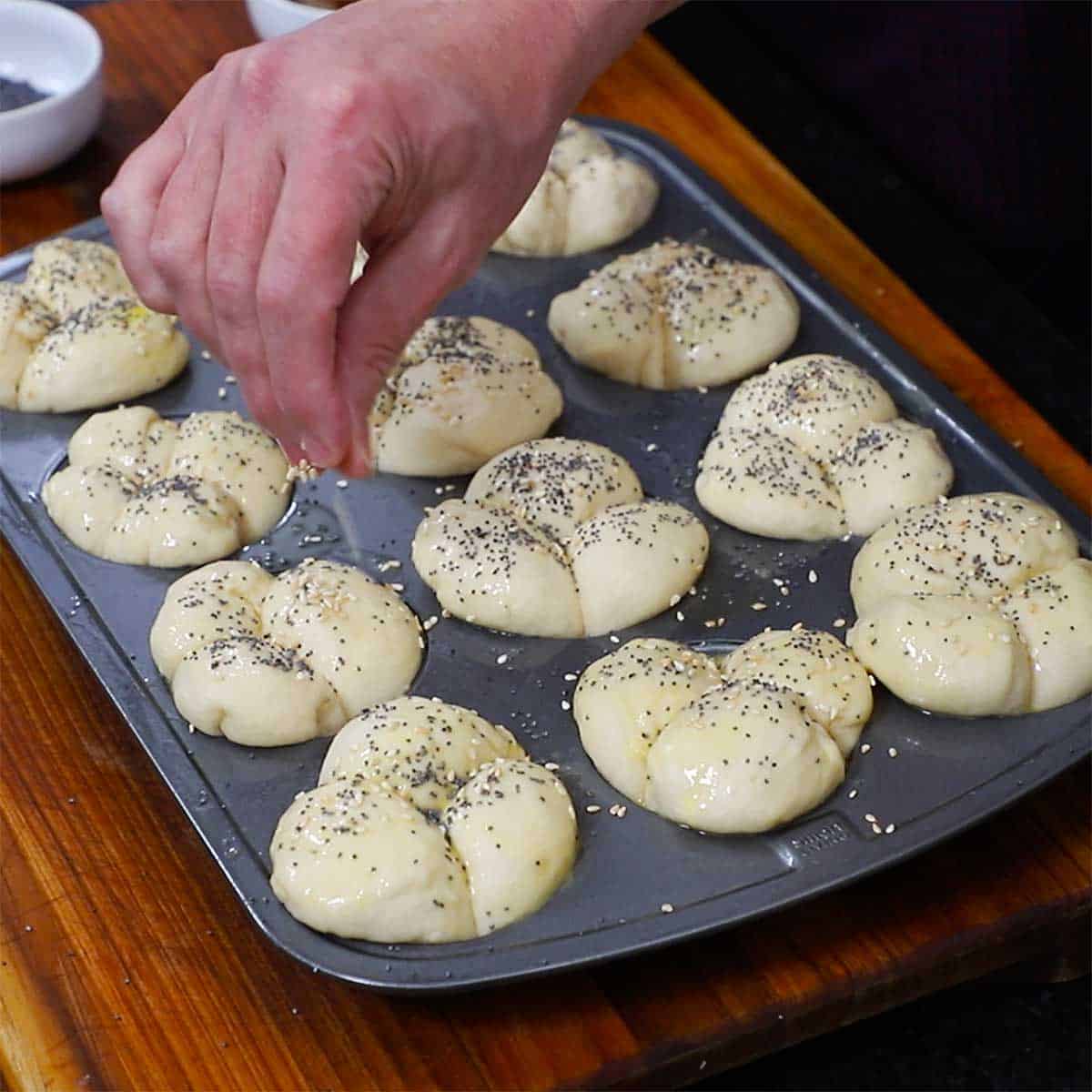 A person sprinkling unbaked cloverleaf dinner rolls with poppy seeds and sesame seeds.