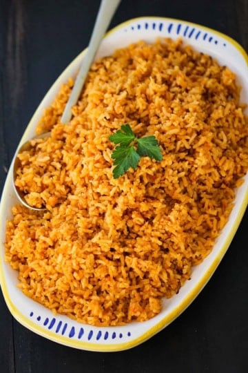 A platter filled with a pile of homemade Mexican rice topped with a sprig of fresh parsley with a large serving spoon inserted in the side.