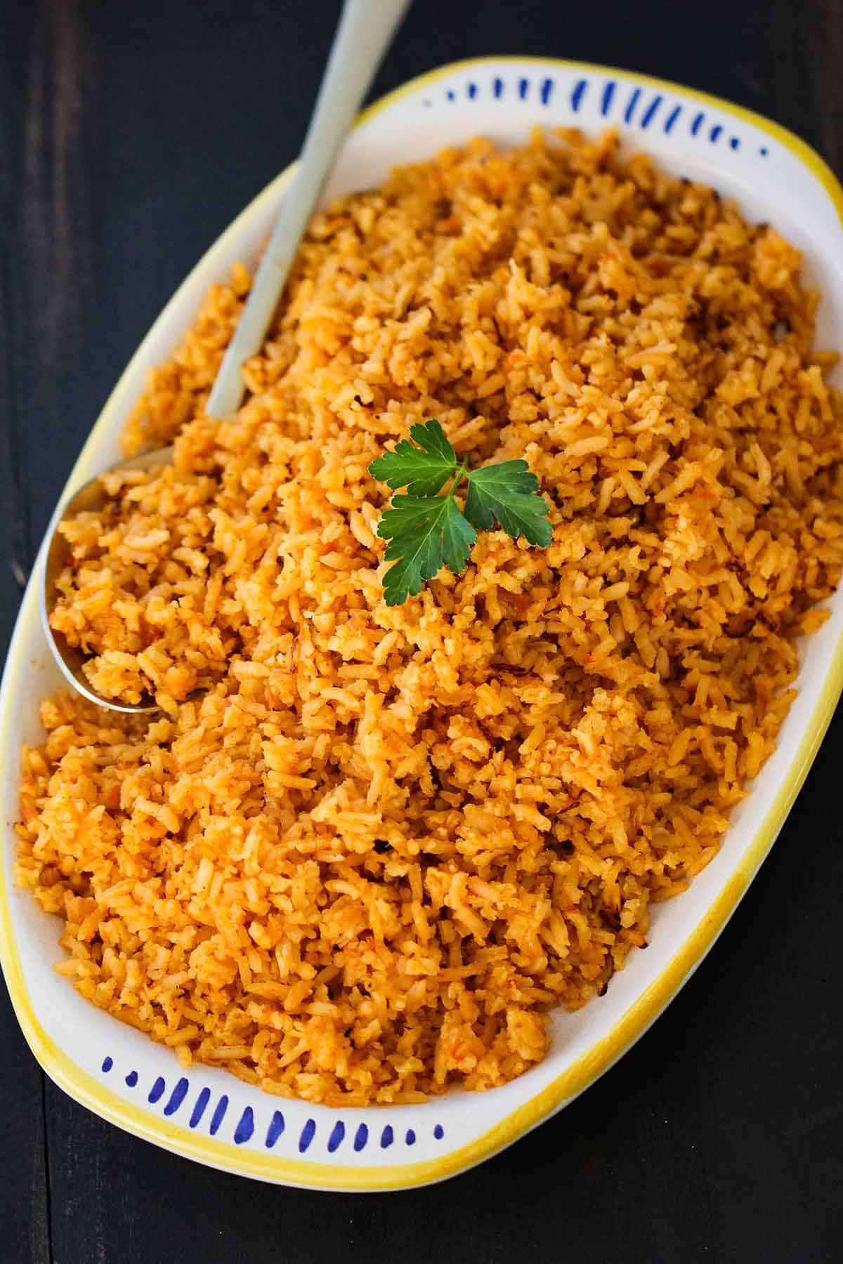 A platter filled with a pile of homemade Mexican rice topped with a sprig of fresh parsley with a large serving spoon inserted in the side.