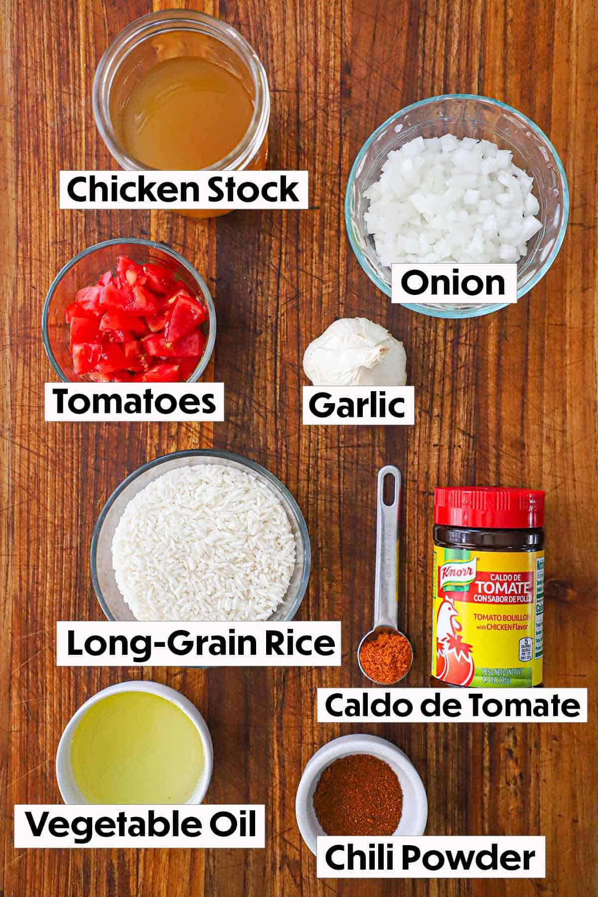 An arrangement of ingredients for Mexican rice on a wooden background including chicken stock, diced onion, chopped tomatoes, garlic, rice, caldo de tomate, oil, and chili powder.