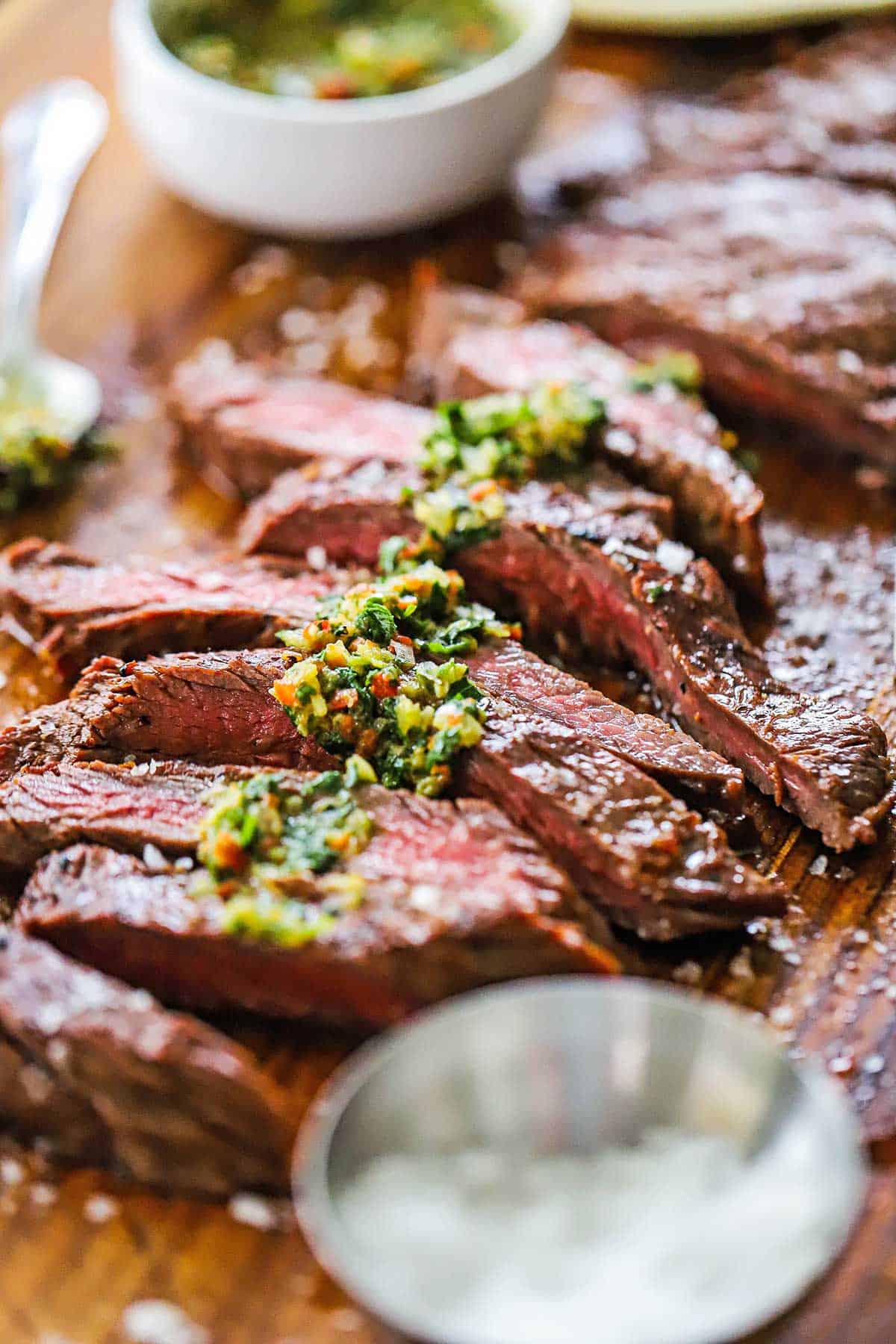 Strips of grilled carne asada that has been cooked to medium-rare and is resting on a cutting board and topped with chimichurri sauce and coarse sea salt.