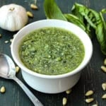 A bowl of easy pesto sauce in a small white bowl next to scattered pine nuts, a head of garlic, and fresh basil leaves.