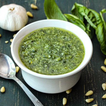 A bowl of easy pesto sauce in a small white bowl next to scattered pine nuts, a head of garlic, and fresh basil leaves.