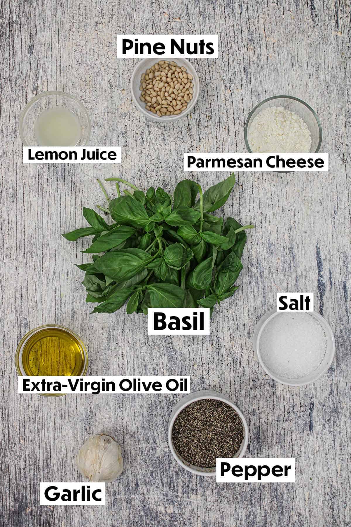 An arrangement of ingredients on a white wooden background including pine nuts, lemon juice, Parmesan cheese, fresh basil, olive oil, salt, pepper, and garlic.