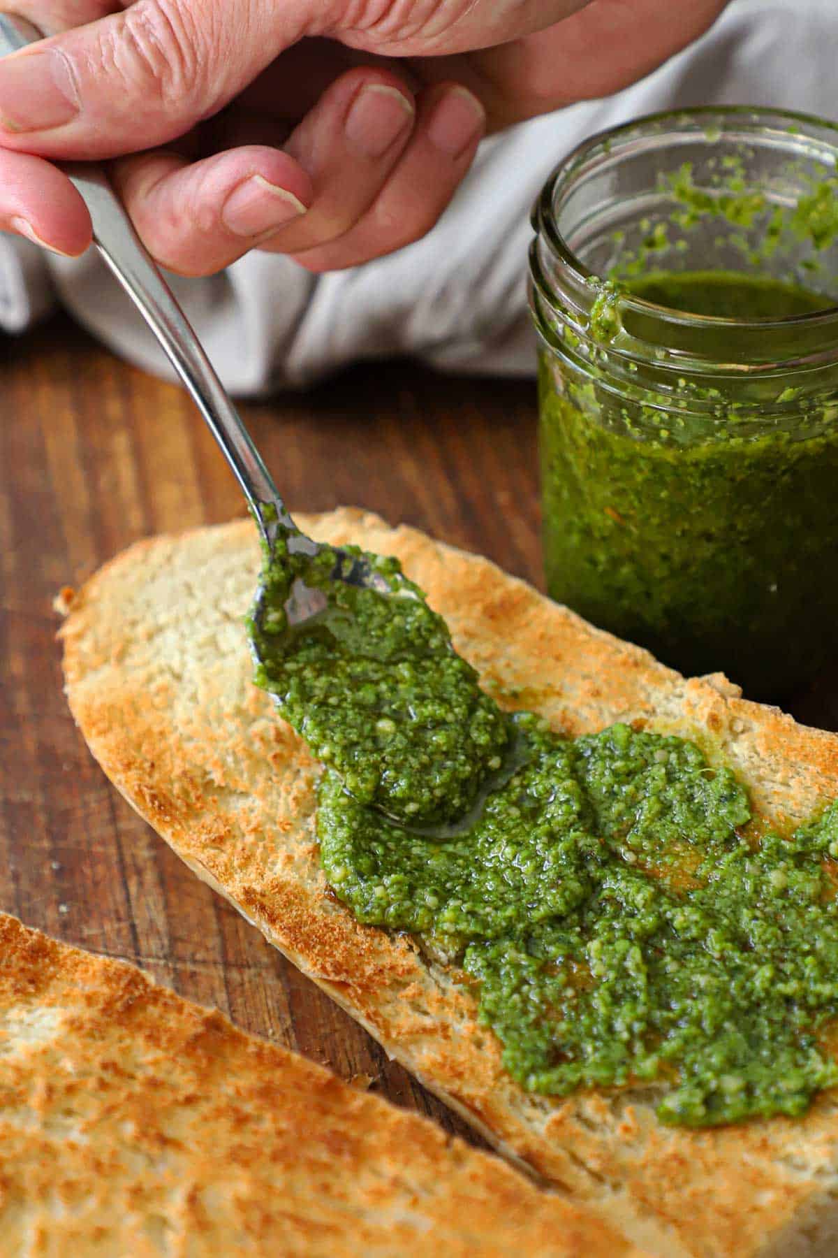 A person using a spoon to spread easy pesto sauce over toasted French bread on a cutting board with a jar of the sauce nearby.