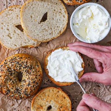 A person spreading cream cheese over the surface of a halved homemade everything bagel on a piece of brown paper next to other bagels and a bowl of cream cheese.