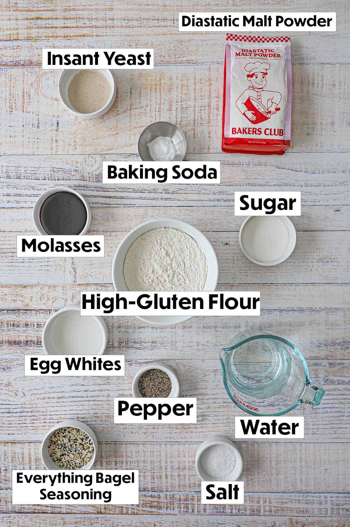 An arrangement of ingredients for homemade everything bagels on a white background including yeast, malt powder, baking soda, sugar, flour, molasses, eggs, salt, pepper, bagel seasoning, and a cup of water.